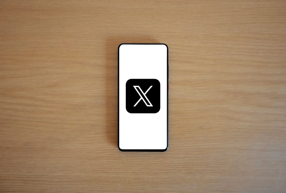 Smartphone showing the X app logo on a wooden desk, illustrating why the European Commission is fining X