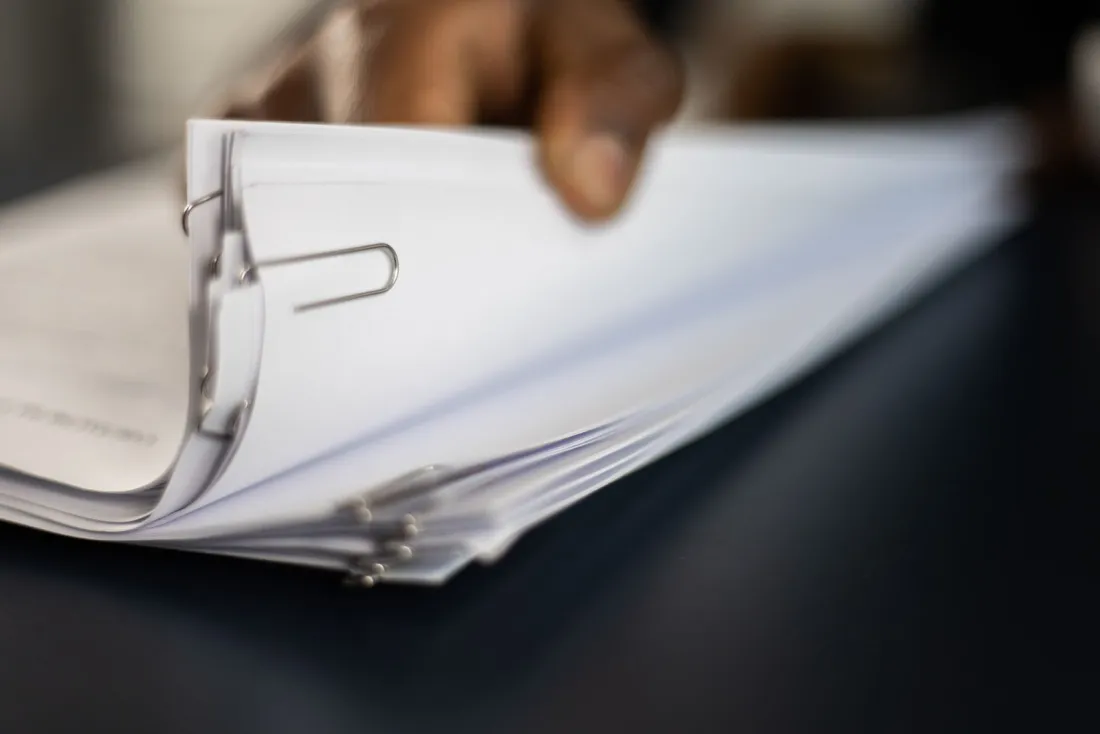 Hand turning a thick stack of clipped transaction documents, illustrating the financing behind the Paramount Warner Bros merger and Fitch's downgrade