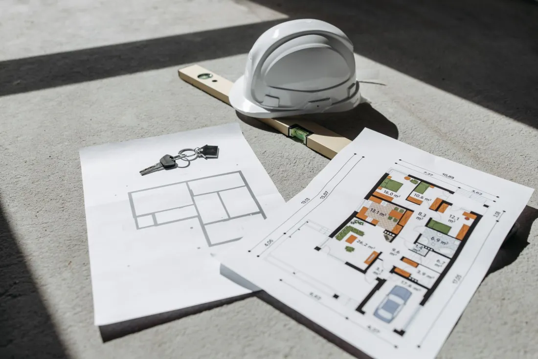House plans, keys and a hard hat on a concrete floor, illustrating what happens if a house builder goes bust before completion