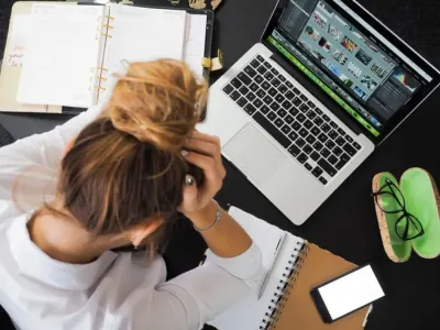 A stressed woman holding her head at a cluttered desk, illustrating emotional abuse at work