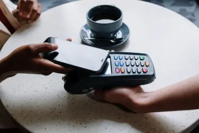 A hand tapping a phone against a card payment terminal at a cafe table, illustrating payment technology