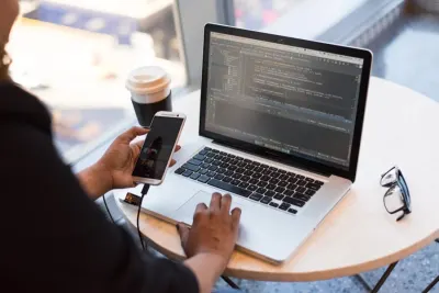 A person checking a smartphone beside a laptop showing code at a cafe table, illustrating employee engagement during lockdown