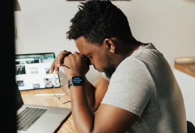 A man resting his head on his hands at a desk in front of a laptop, illustrating increasingly demanding working lives