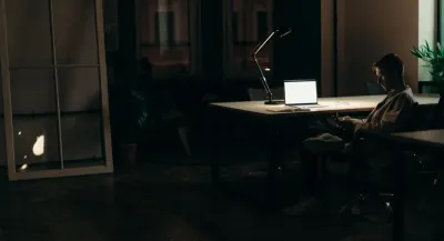 A man working alone at a desk lit by a lamp in a dark office at night, reflecting a pivotal stage for the future of work