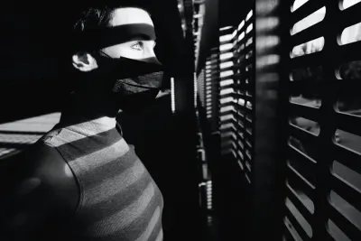 A black and white portrait of a person wearing a face mask looking out through window blinds, reflecting COVID's impact on careers