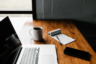 Tidy wooden desk with a coffee cup, notebook and phone beside an open laptop, illustrating hybrid working for companies