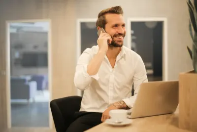 Smiling businessman on the phone with a laptop and coffee at his desk, illustrating job satisfaction